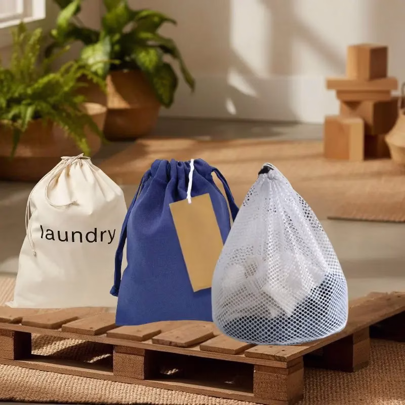 Collection of eco-friendly laundry bags made from organic cotton (tan) and recycled PET fabric (earth tones), displayed on a wooden surface with small certification badges or QR codes visible